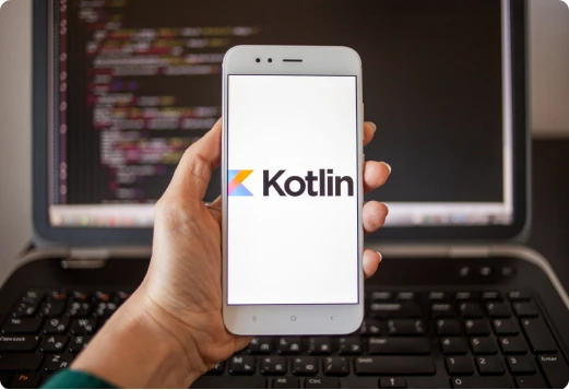 A phone showing the Kotlin logo, held in front of a laptop with code on screen