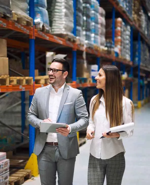 Two colleagues walking a warehouse aisle with a tablet and clipboard