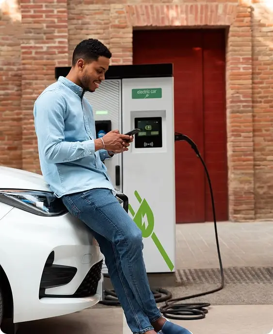 A man sitting on the bonnet of a white electric car, phone in hand, while it charges from a public charge point against a brick wall