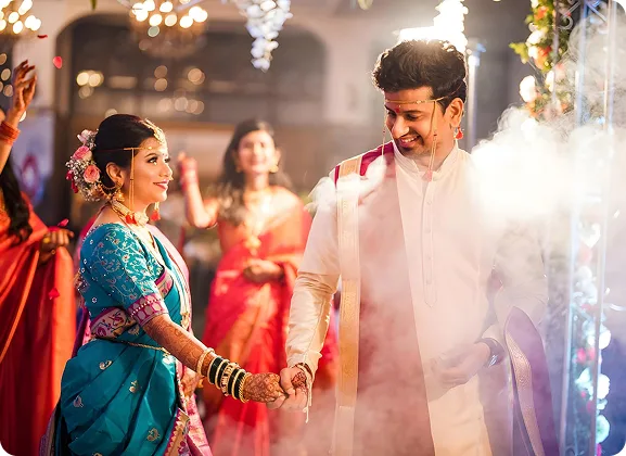 A bride and groom in traditional Maharashtrian dress holding hands at their wedding, guests behind them