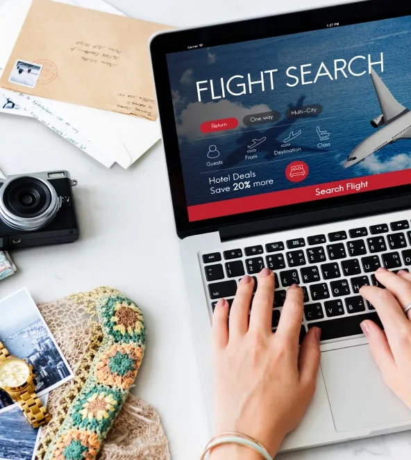 Someone searching for flights on a laptop, beside a camera and printed photographs
